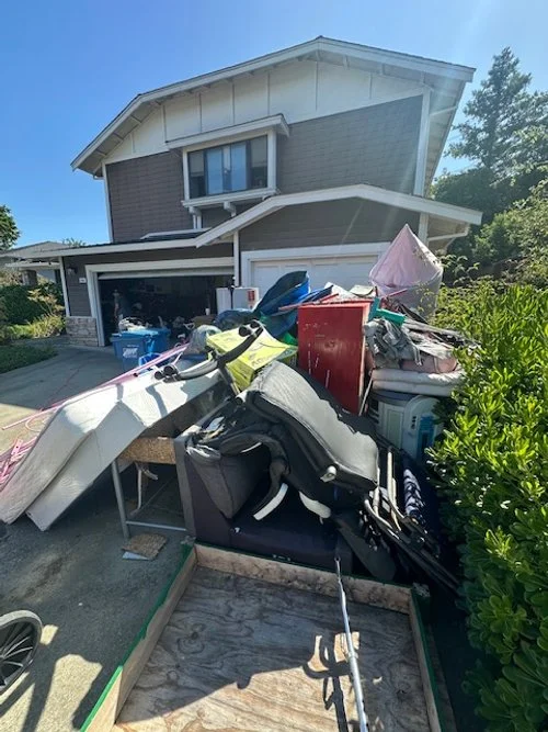 Driveway cluttered with various items including furniture, a golf bag, and household belongings in front of a two-story house with a garage door.