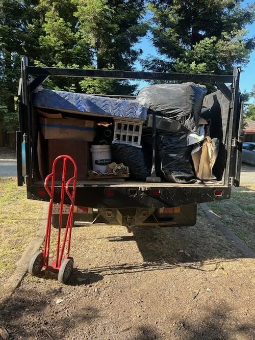 The image shows the back of a black truck loaded with various items, including a blue mattress, black trash bags, cardboard boxes, and miscellaneous household belongings. A red hand truck is parked beside the truck on the left side. The scene is outdoors with green trees and a partly cloudy sky in the background.