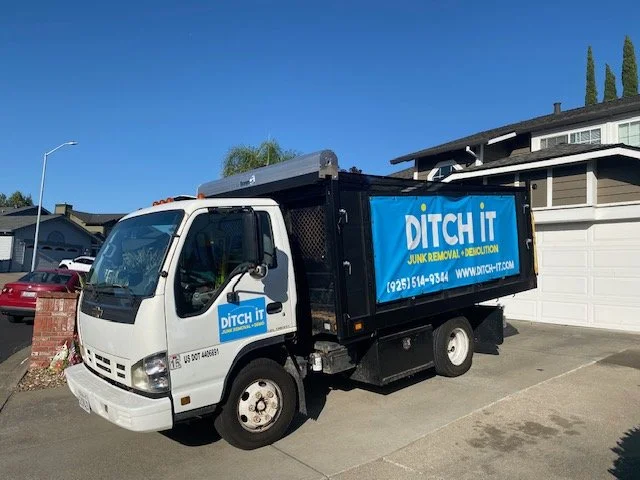 A small white trash removal truck parked in a driveway, with a blue banner on the side reading 'Ditch It Junk Removal & Demolition' and contact information, in front of a suburban house with a gray roof.