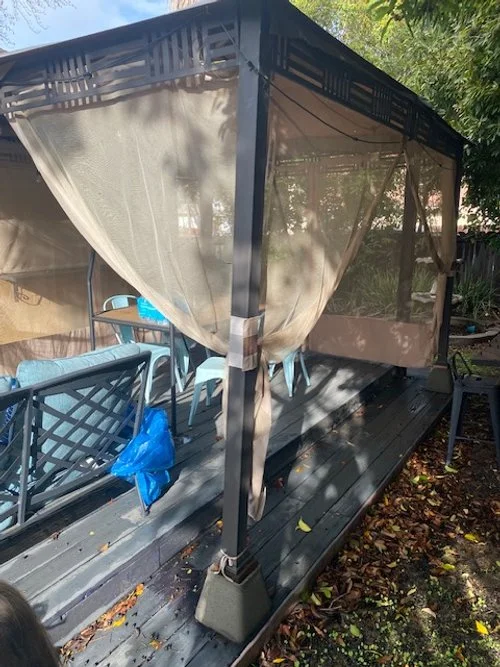 Outdoor gazebo with fabric curtains and a deck, featuring patio furniture and surrounded by trees.