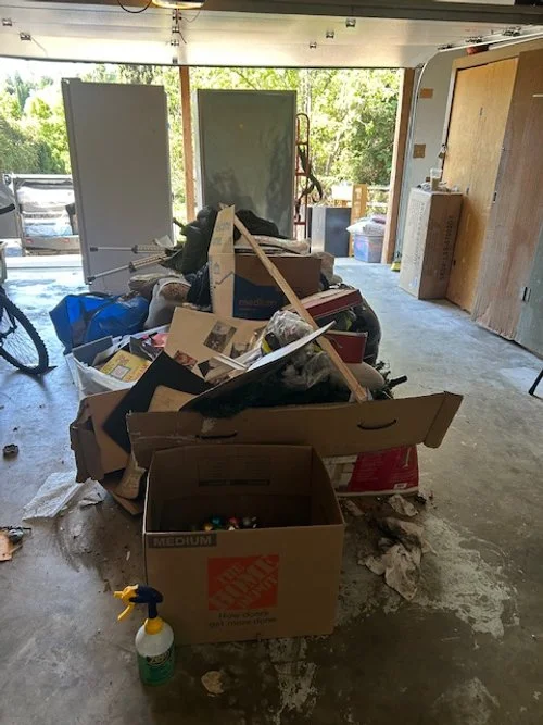 Cluttered garage with cardboard boxes, miscellaneous items, and debris.