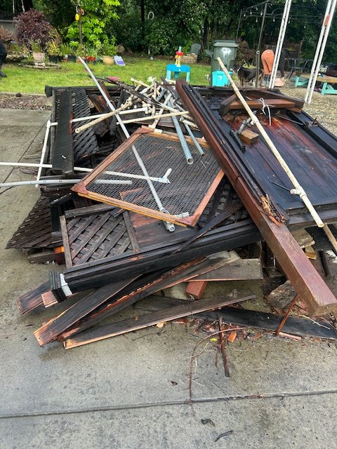 Pile of broken wooden outdoor furniture, metal poles, and glass window pieces on a concrete surface in a backyard.