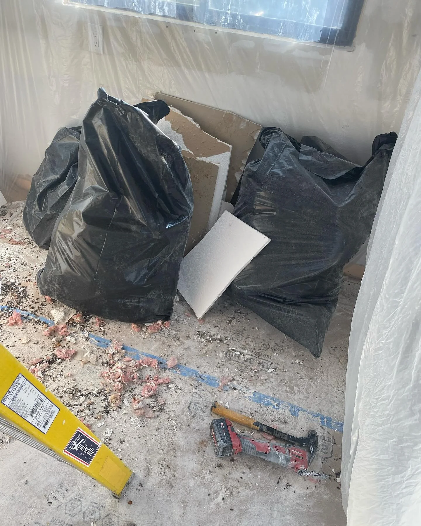 Construction debris with two black garbage bags, pieces of drywall, a cordless power drill, a yellow level, and insulation material on the floor.