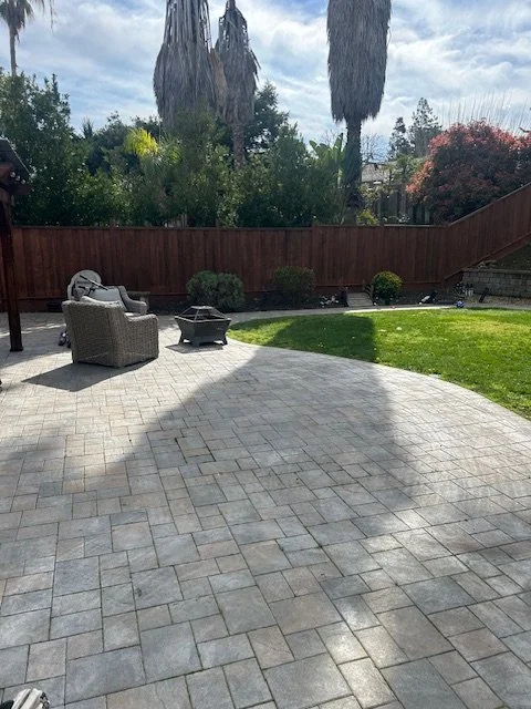 Backyard patio with outdoor furniture, a fire pit, grass, trees, and a wooden fence.