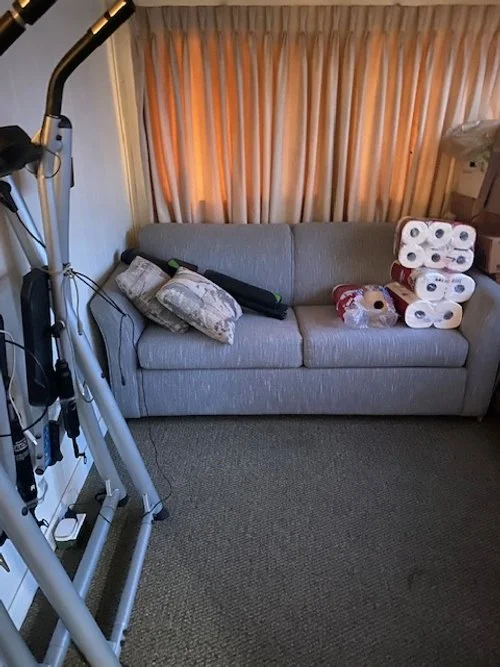 A gray sofa with decorative pillows and stacked paper towels, next to exercise equipment, in a room with carpeted floor and curtains.