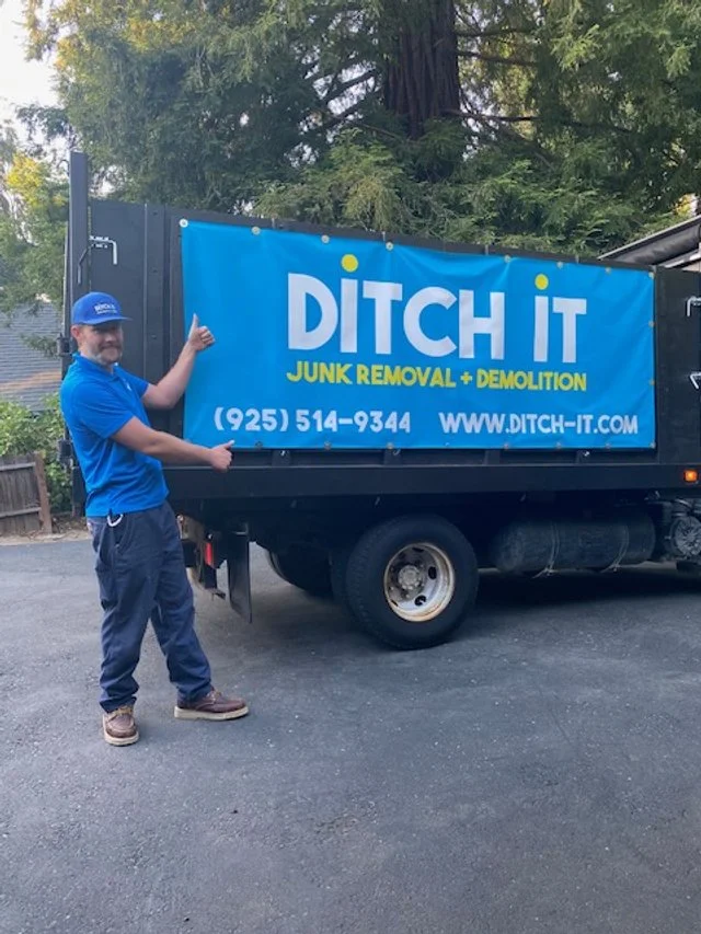 A man in a blue uniform and cap standing next to a black truck with a blue banner that reads "DITCH IT Junk Removal + Demolition," along with a phone number and website.