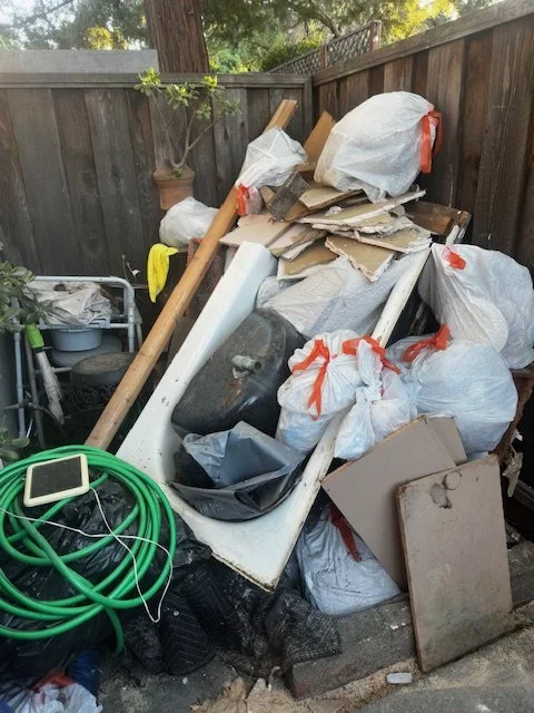 Piles of assorted trash including cardboard, plastic bags, a bathroom sink, and gardening tools in a backyard corner surrounded by a wooden fence.