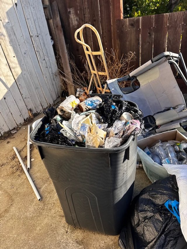 A large black trash can filled with burnt garbage, surrounded by discarded household items and a wooden crutch leaning against it, outdoors near a wooden fence.