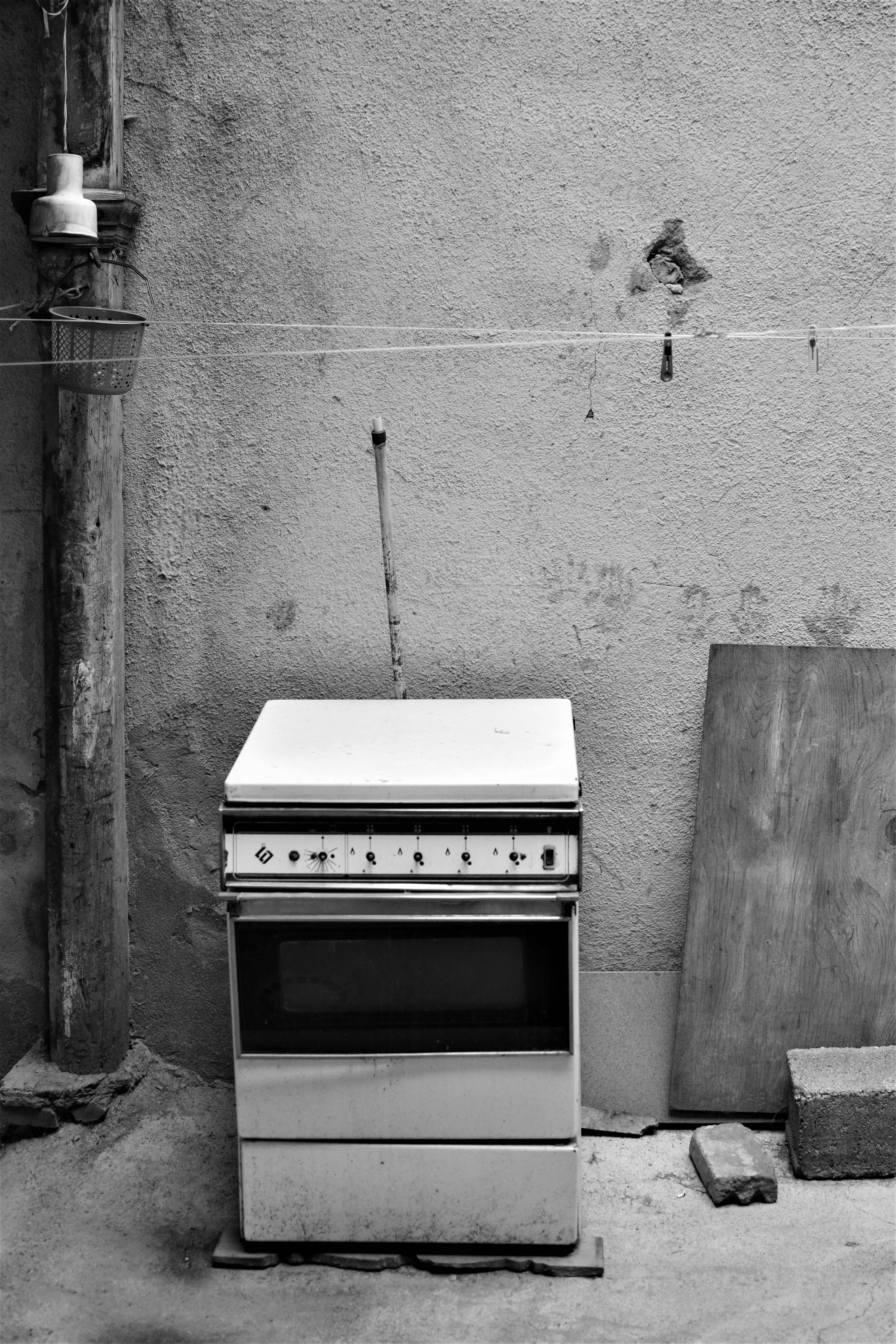 Black and white image of an old stove against a wall, with a wooden plank and bricks nearby, and a hanging basket on a pole.