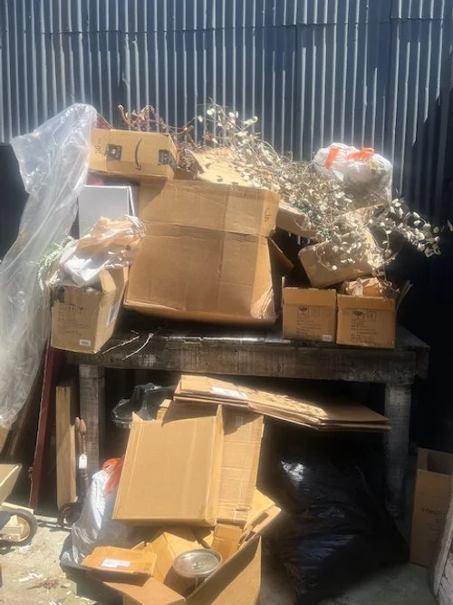 Piles of cardboard boxes and packaging materials scattered on and around a wooden workbench with a corrugated metal fence in the background.