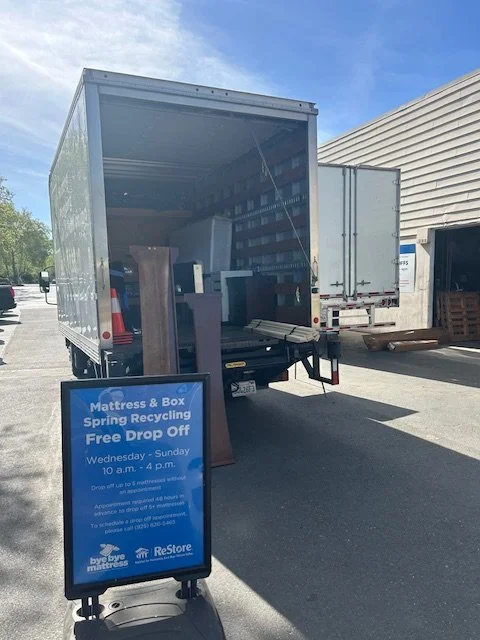 Open delivery truck with furniture inside, parked near a building with a signboard advertising mattress recycling and free drop-off, on a sunny day.