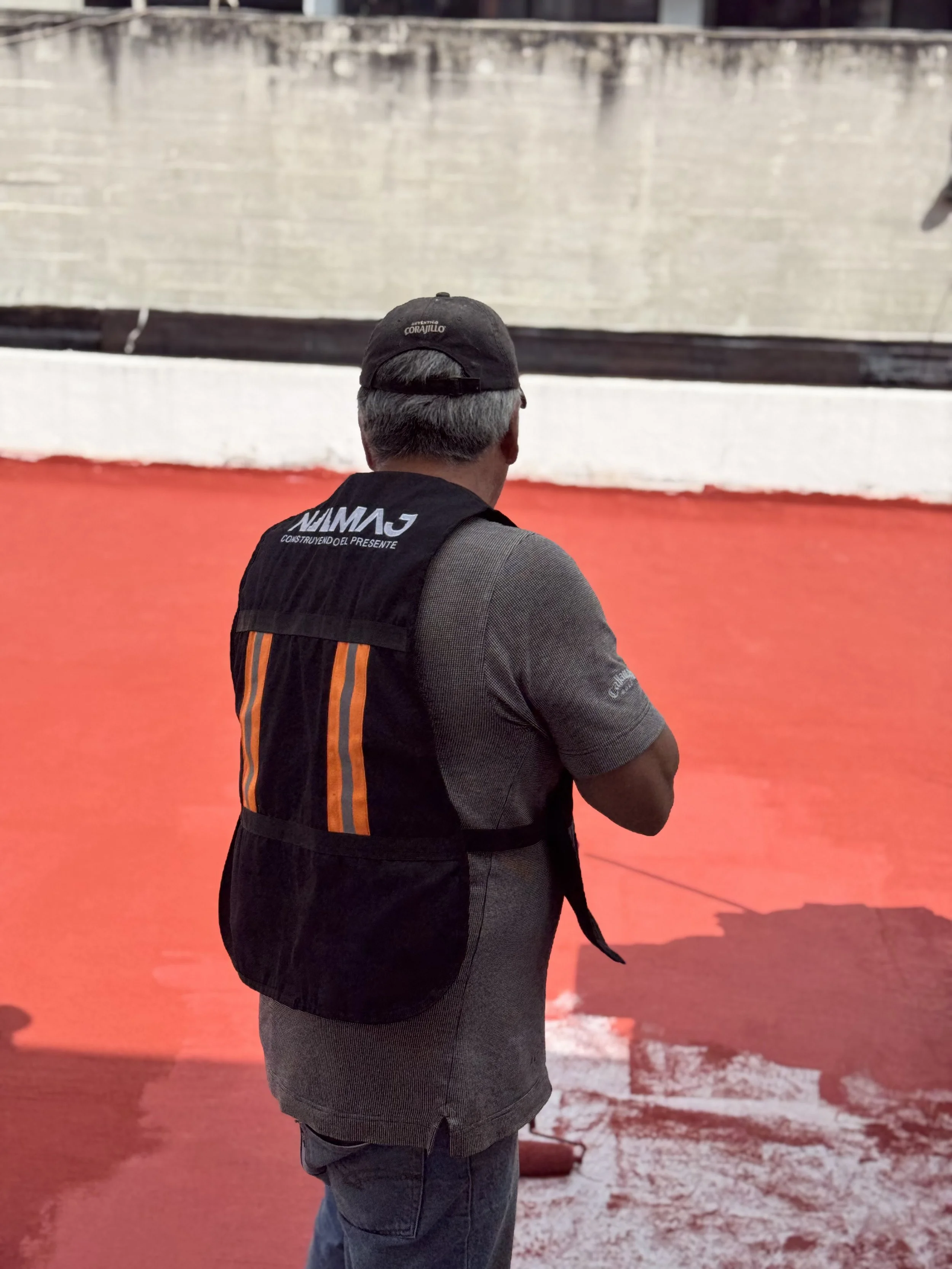Hombre con gorra y chaqueta, de espaldas, sosteniendo un cable en una superficie roja, con muro de fondo.
