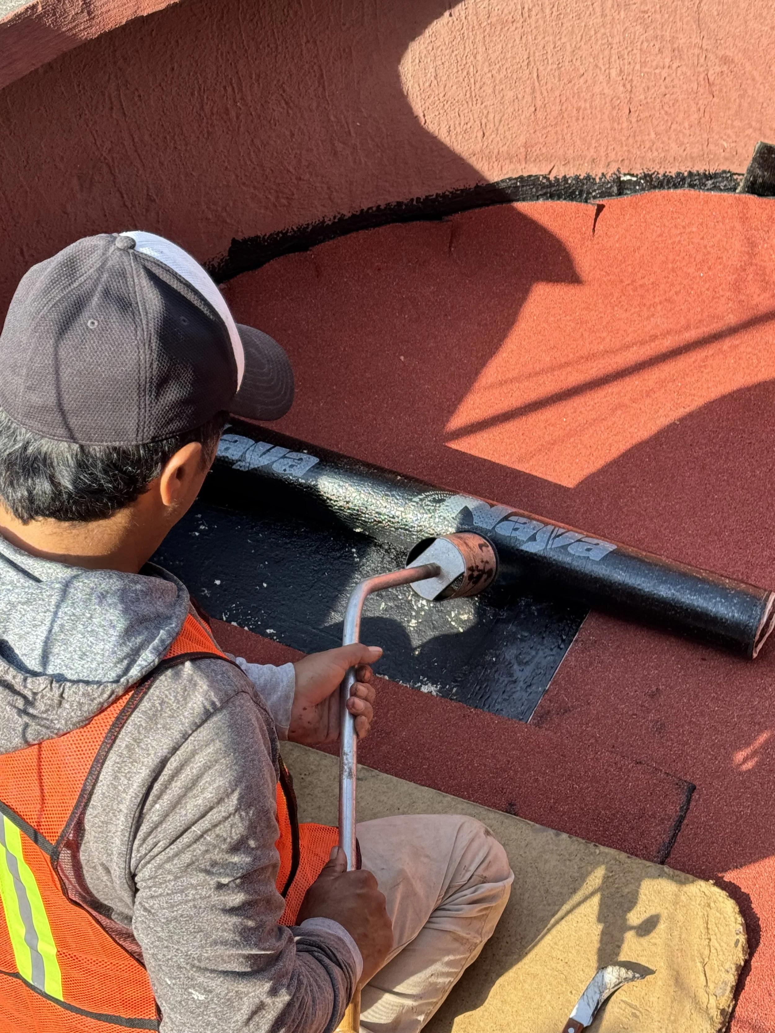 Hombre en un chaleco de seguridad aplicando impermeabilizante prefabricado con un soplete sobre una superficie roja, en un espacio cubierto con paredes de color terracota y sombra proyectada.