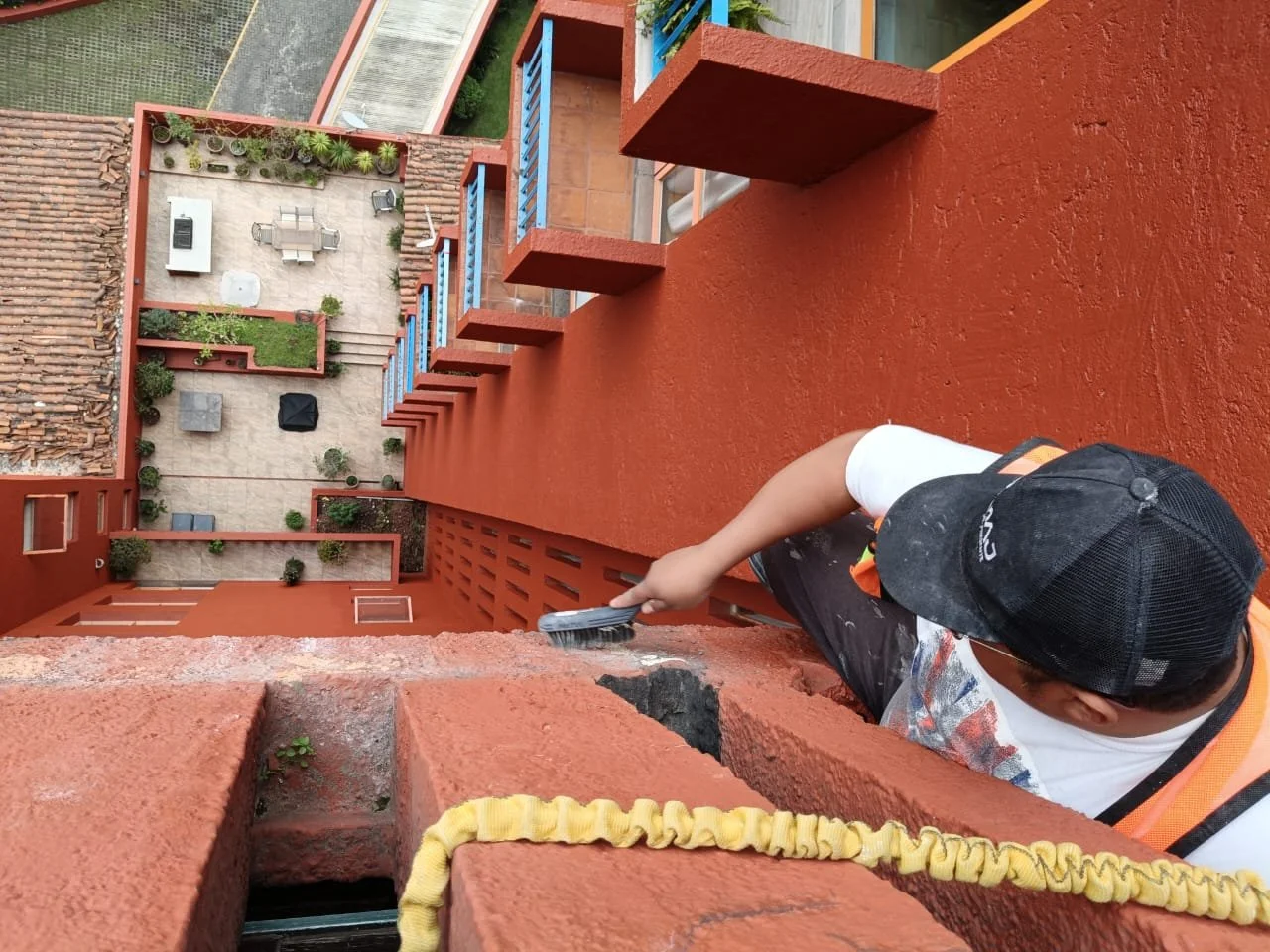 Persona limpiando la pared de un edificio alto con brocha, vista desde arriba, con terraza y áreas verdes abajo.
