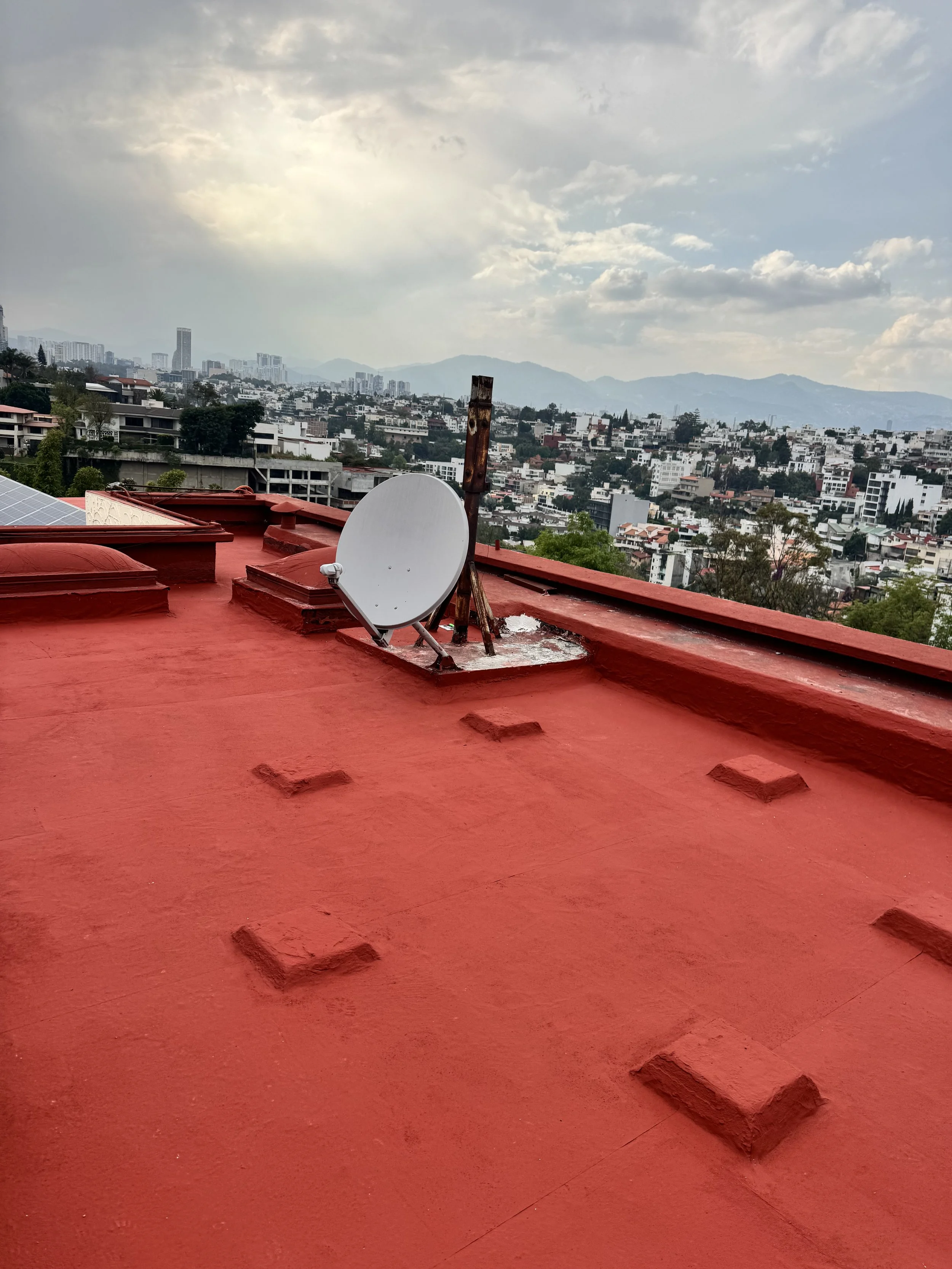 Techo de azotea rojo con antena parabólica y vista de la ciudad con edificios y montañas al fondo bajo cielo nublado.