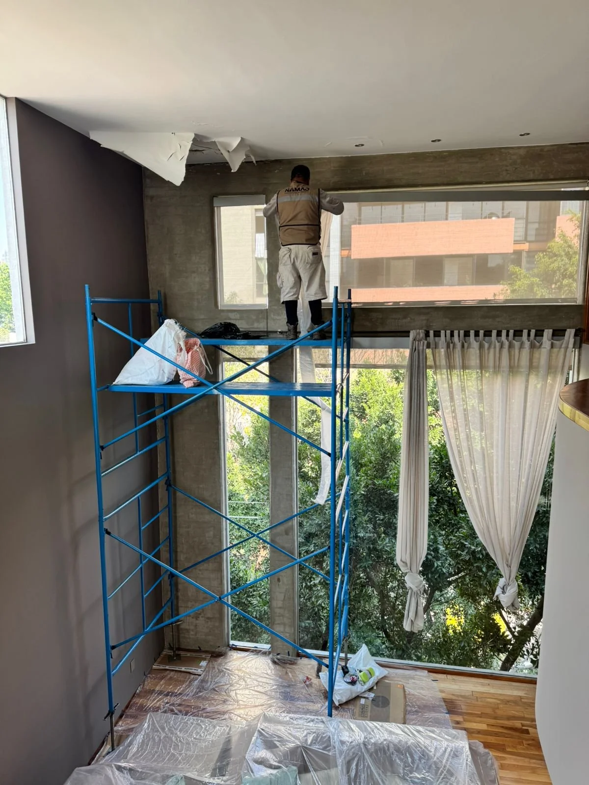 Un trabajador de construcción con chaleco y pantalones claros en un andamio azul está reparando o pintando el techo de una habitación grande con ventanas amplias y cortinas blancas. La habitación tiene plantas verdes visibles a través de las ventanas