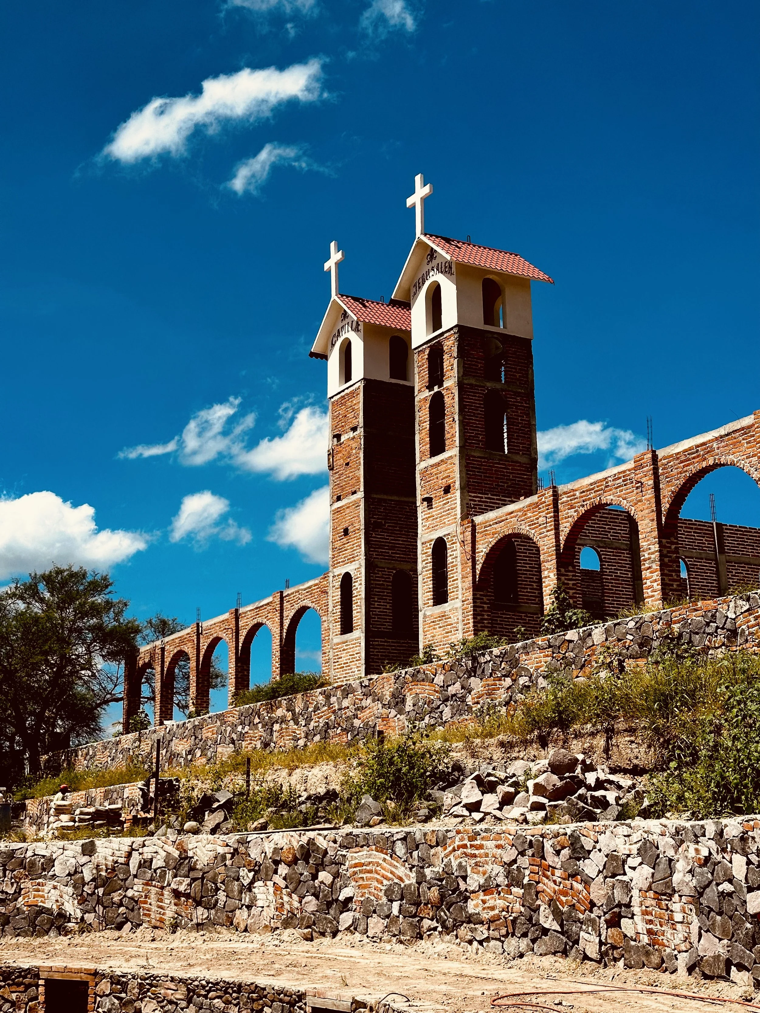 Dos torres de ladrillo con cruces en la cima y arcos en la parte inferior, en un día soleado con cielo azul y algunas nubes, en un lugar con muro de piedra y vegetación.