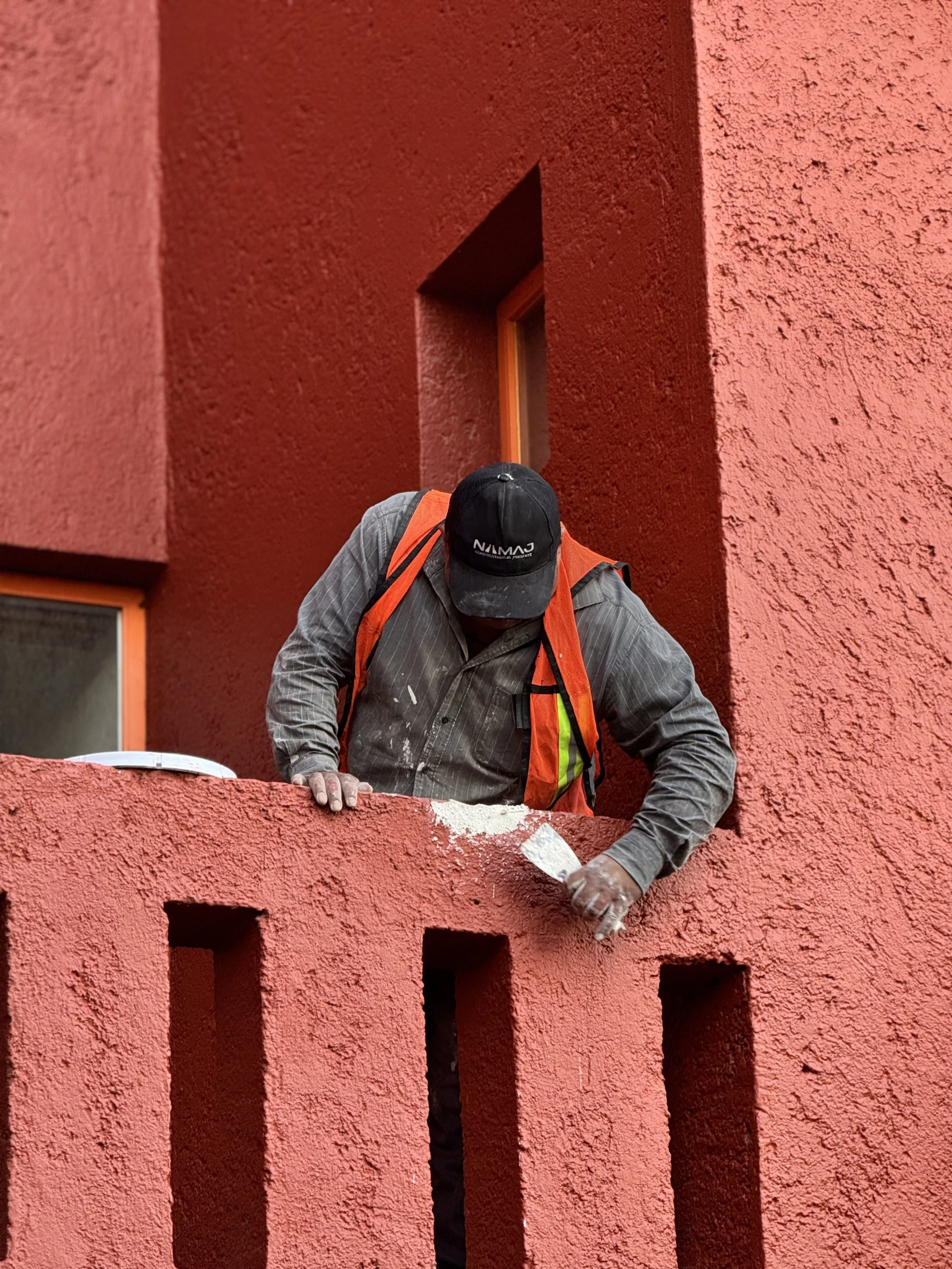 Un albañil pinta la pared de un edificio rojo, usando una camiseta a rayas gris, gorra negra y chaleco naranja, en un balcón con barandal y ventanas también de color rojo.