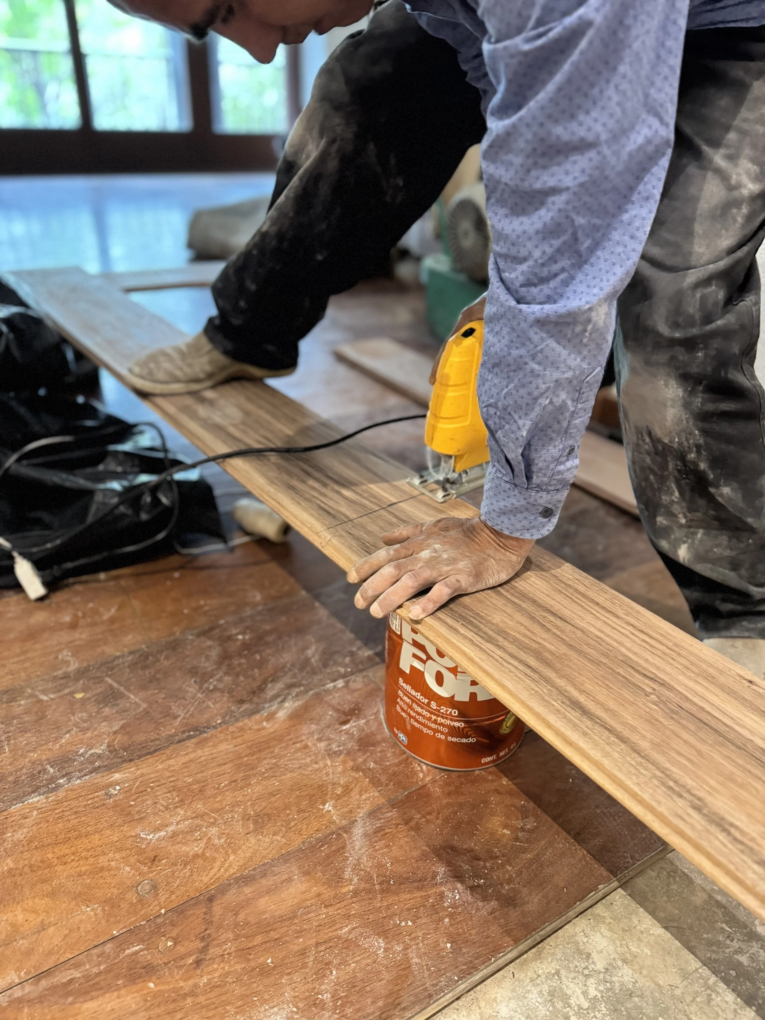 Persona cortando madera con una sierra eléctrica apoyada en un envase de sellador, en un ambiente interior con ventanas y piso de madera, mano sucia y ropa de trabajo.