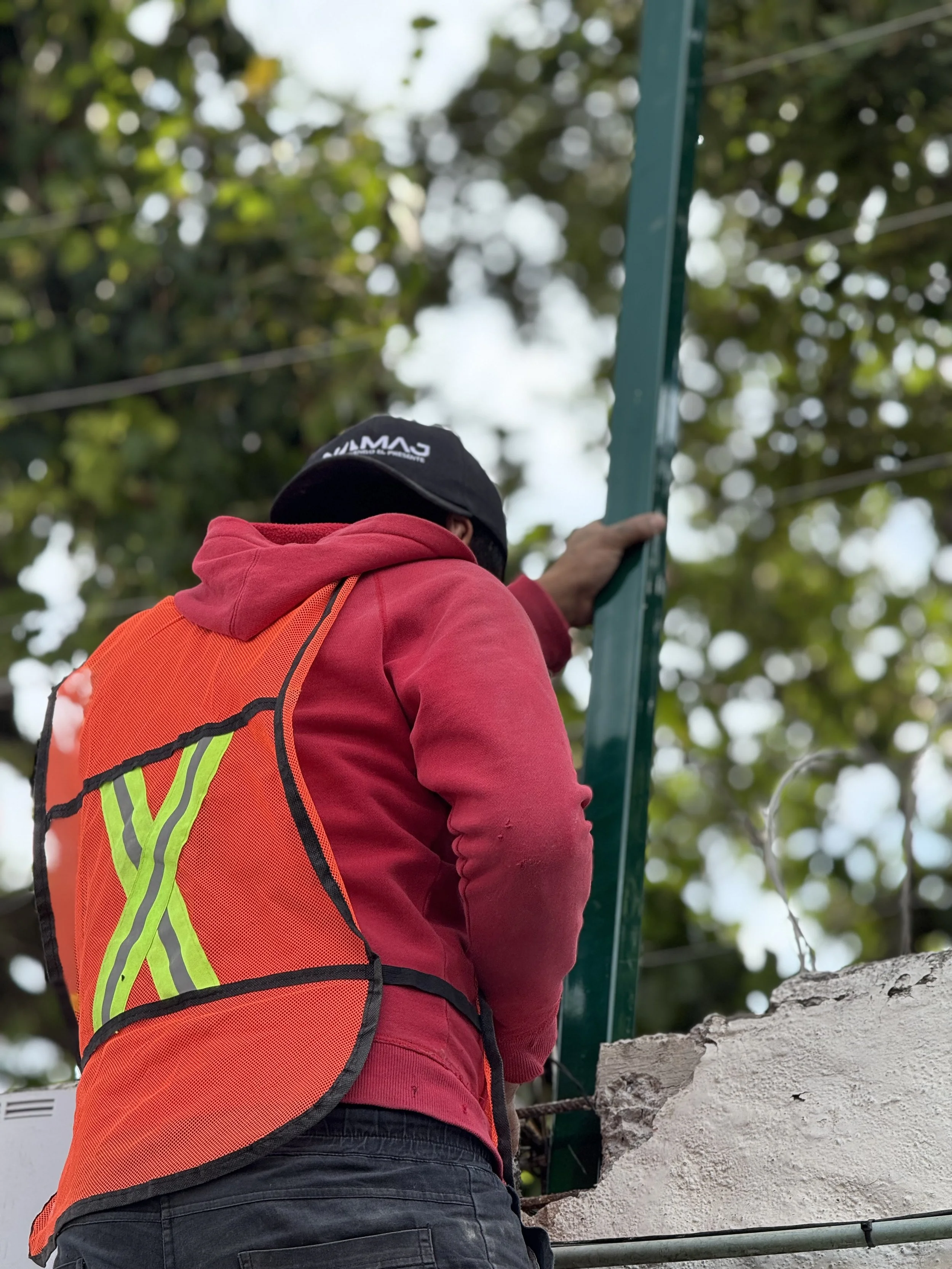 Persona con chaqueta roja y chaleco reflectante naranja trabaja en construcción, sosteniendo una estructura metálica bajo un árbol.