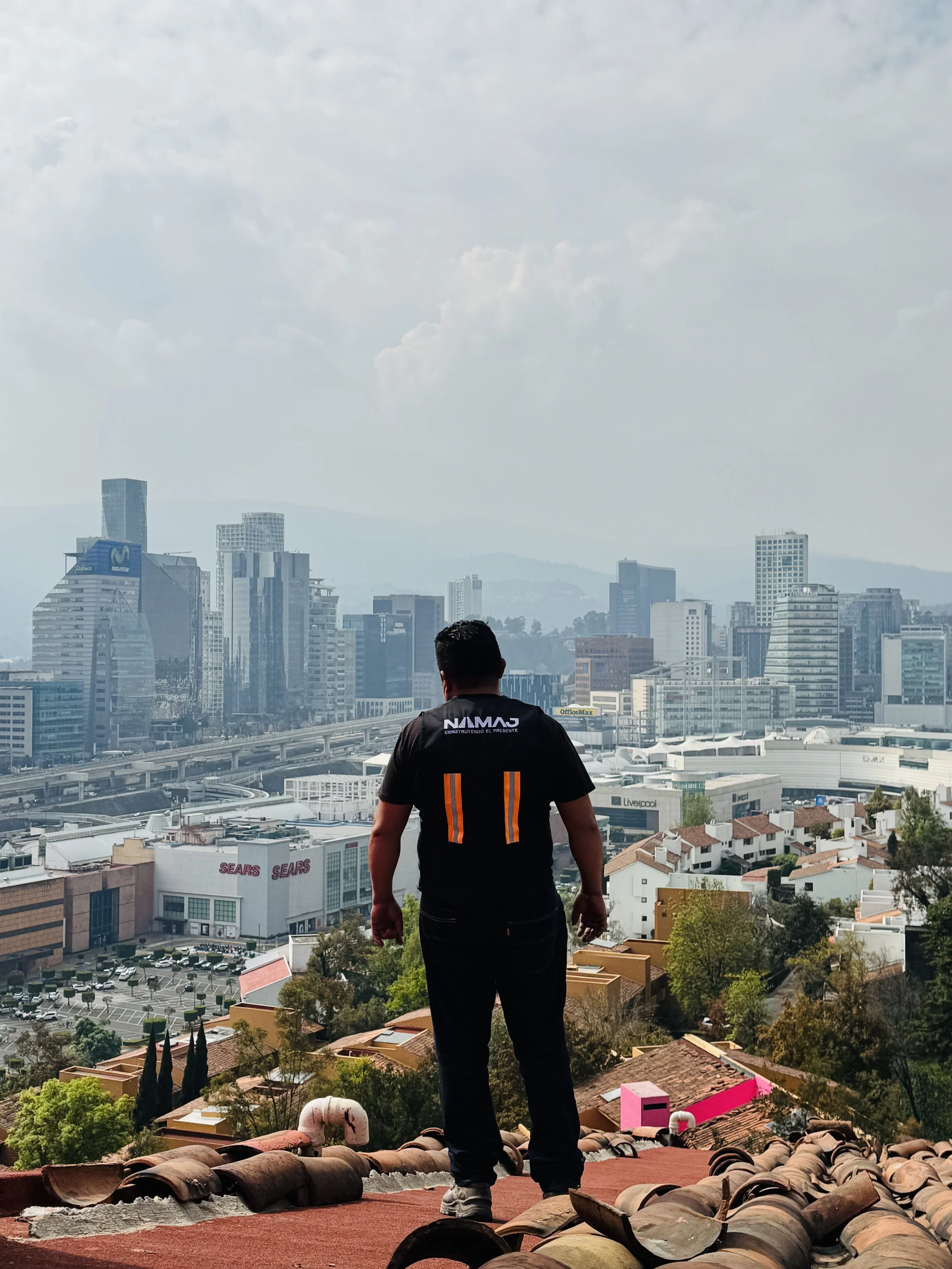 Persona de espaldas de pie en un techo de tejas, mirando hacia una ciudad moderna con edificios altos y un cielo parcialmente nublado.