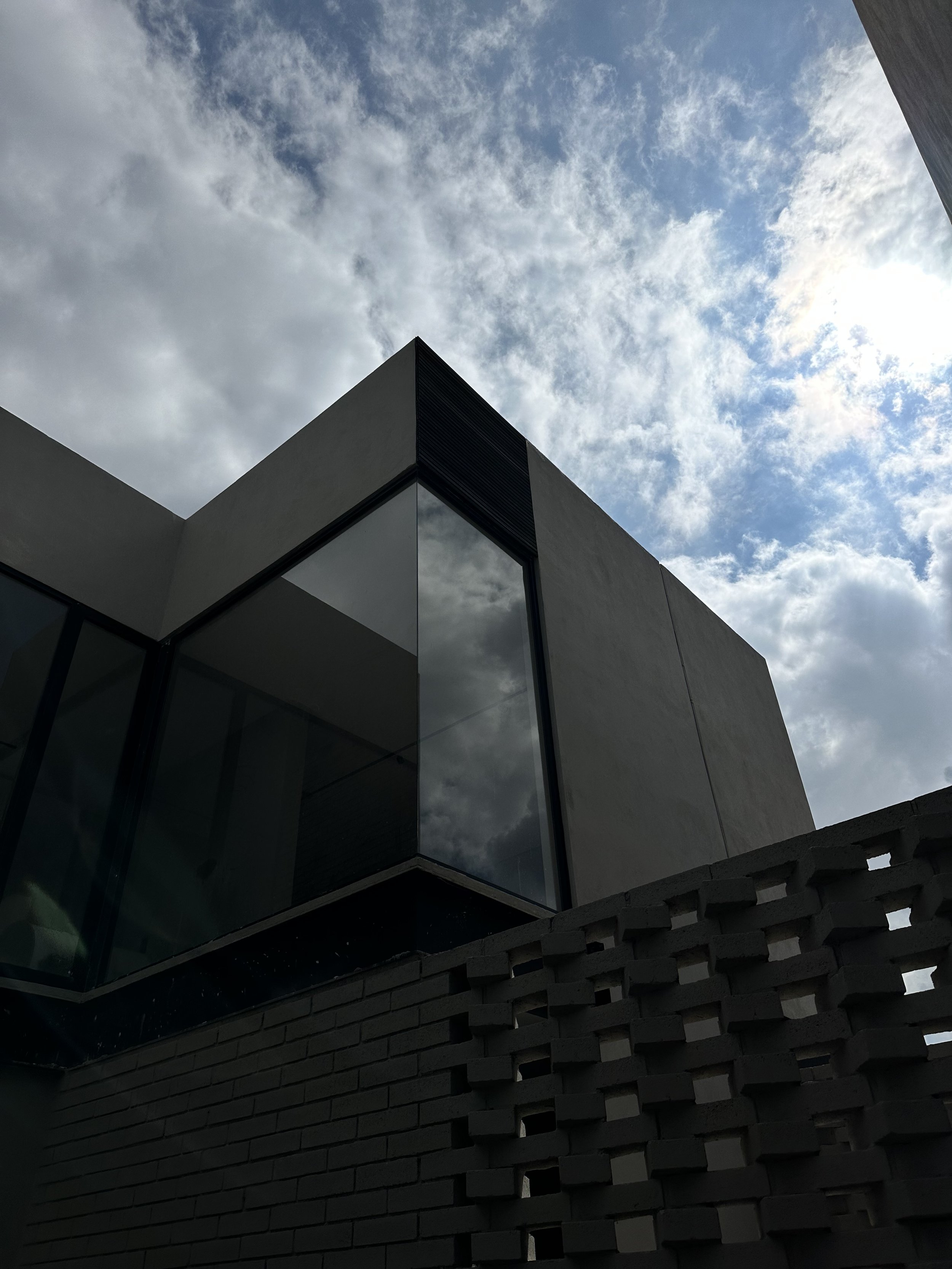 Vista de un edificio moderno con paredes de color oscuro y ventanas de cristal, con cielo nublado y una parte del sol asomándose.