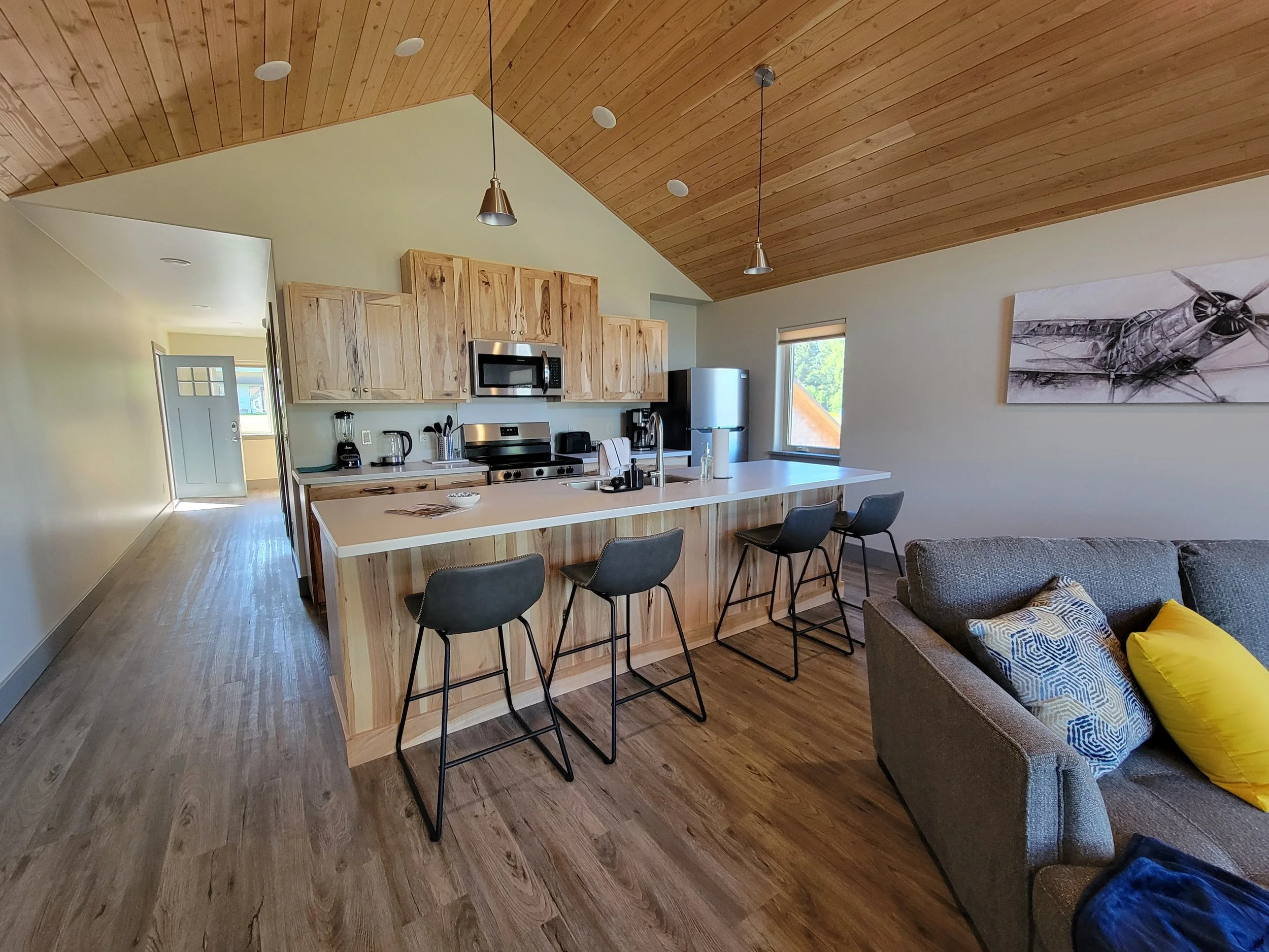 Modern kitchen and living area with wooden ceiling, bar stools, gray sofa, colorful pillows, and appliances.