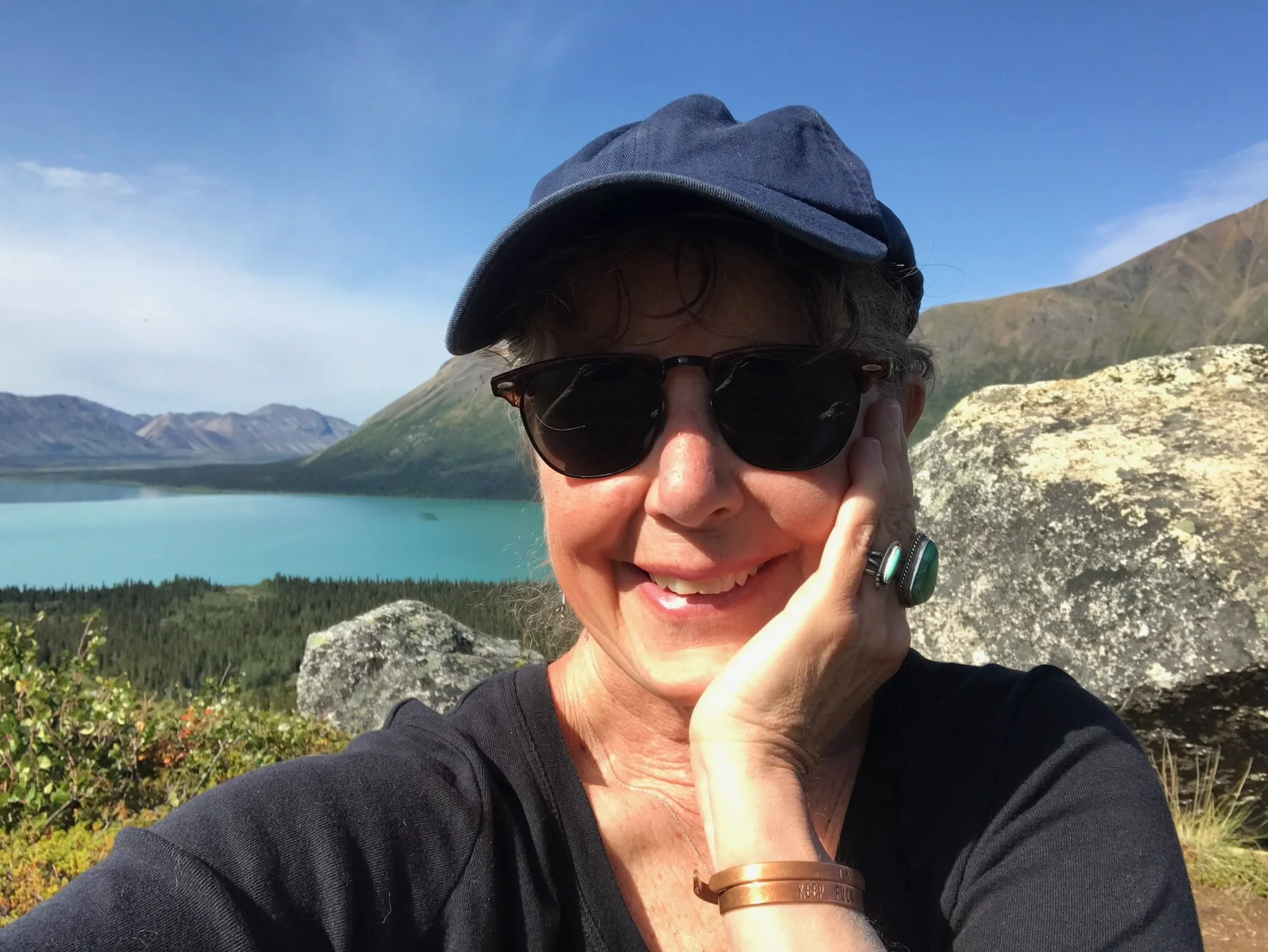 Terrilynn wearing a blue baseball hat and sunglasses smiling in front of a lake.