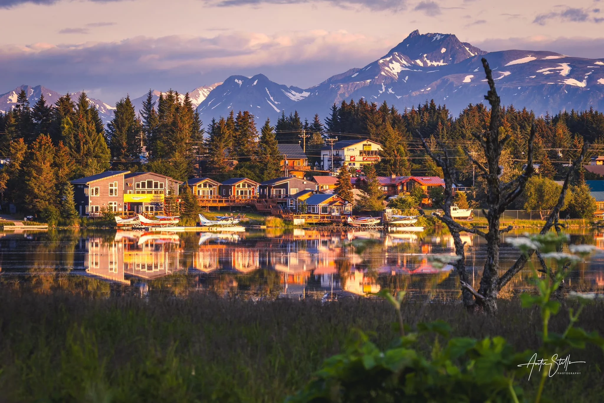 A scenic view of a small town with colorful houses by a reflective lake, surrounded by dense trees and mountains in the background, under a partly cloudy sky at sunset.