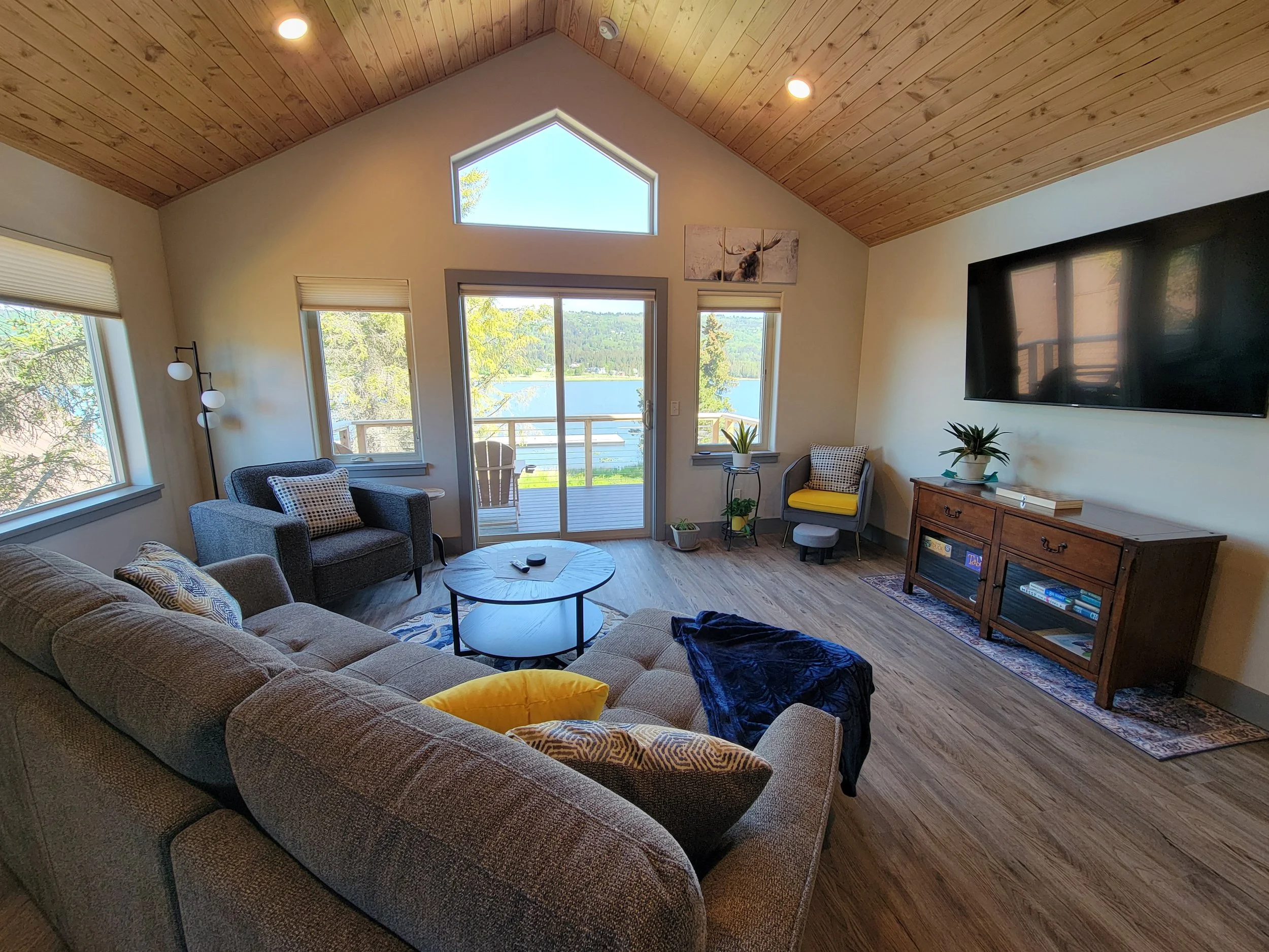 Cozy living room with gray sectional sofa, armchairs, and a large wall-mounted TV. A wooden TV stand holds books and decor. The room features a high wood-paneled ceiling and large windows revealing a scenic outdoor view. A small round coffee table and decorative pillows are present.