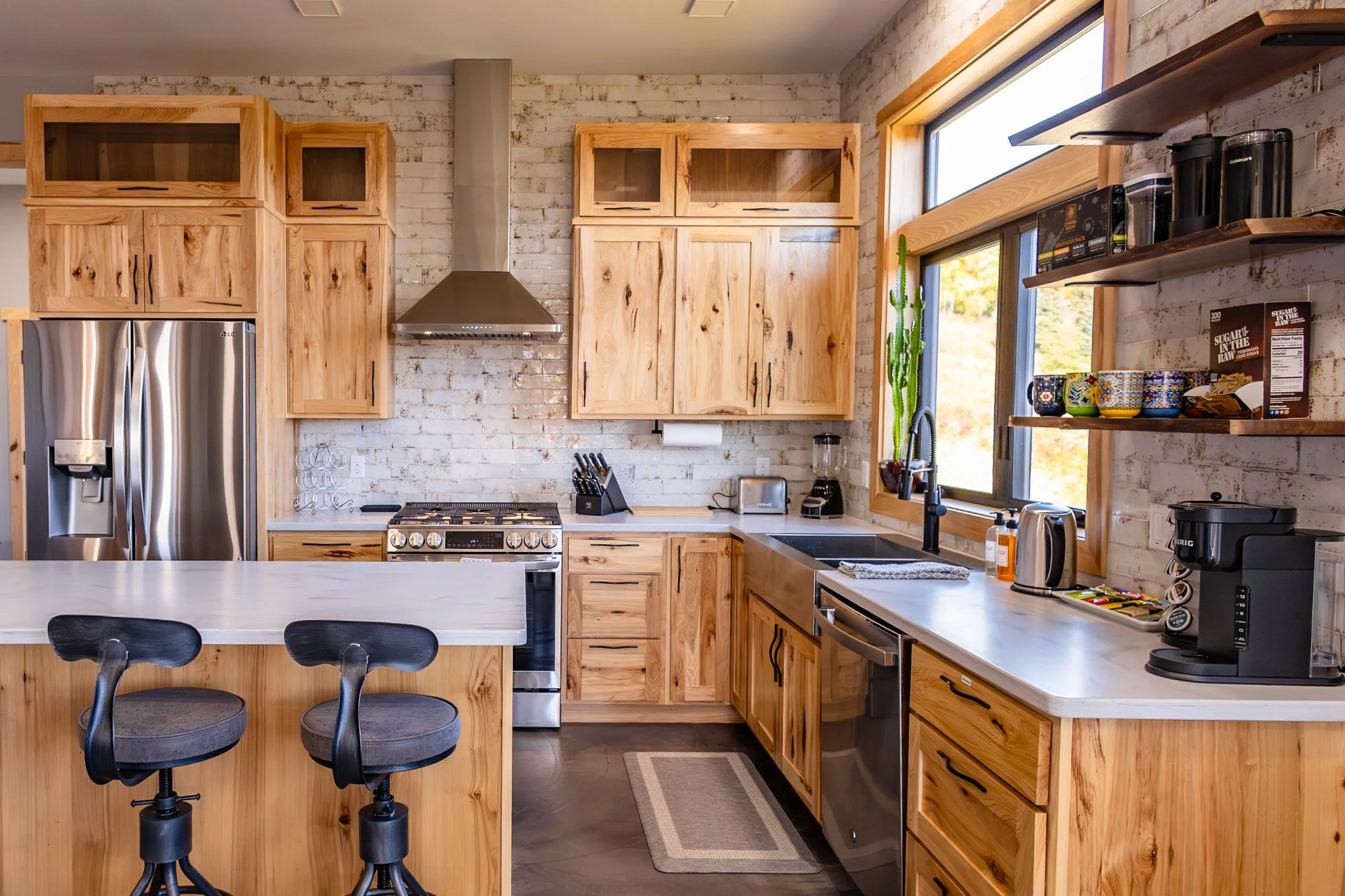 Modern kitchen with stainless steel appliances, wooden cabinets, island with two bar stools, and open shelves displaying mugs and coffee supplies. A large window illuminates the space, and a cactus plant sits by the sink.