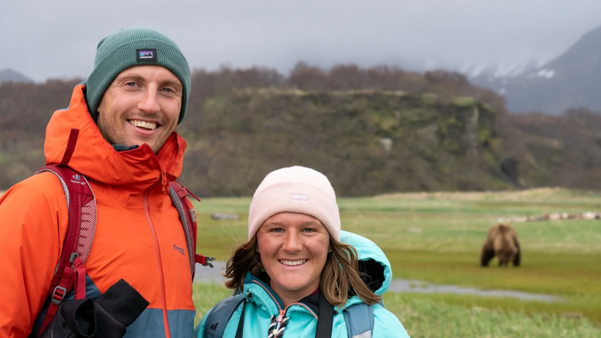 Two guests, man and woman, standing in front of a brown bear at Katmai National Park with Emerald Air Service