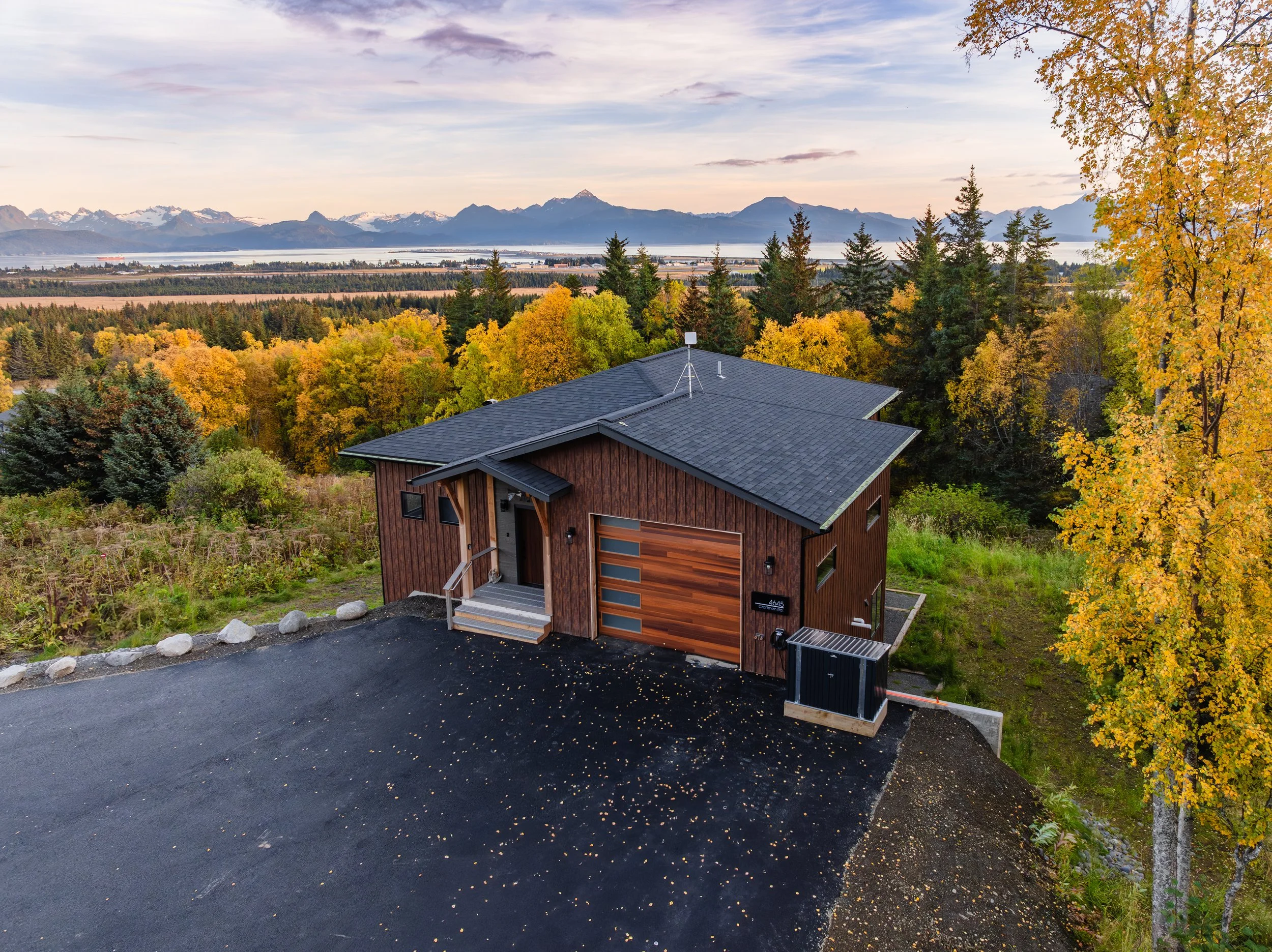 Modern house with wooden exterior surrounded by autumn trees, mountain range and body of water in the background.