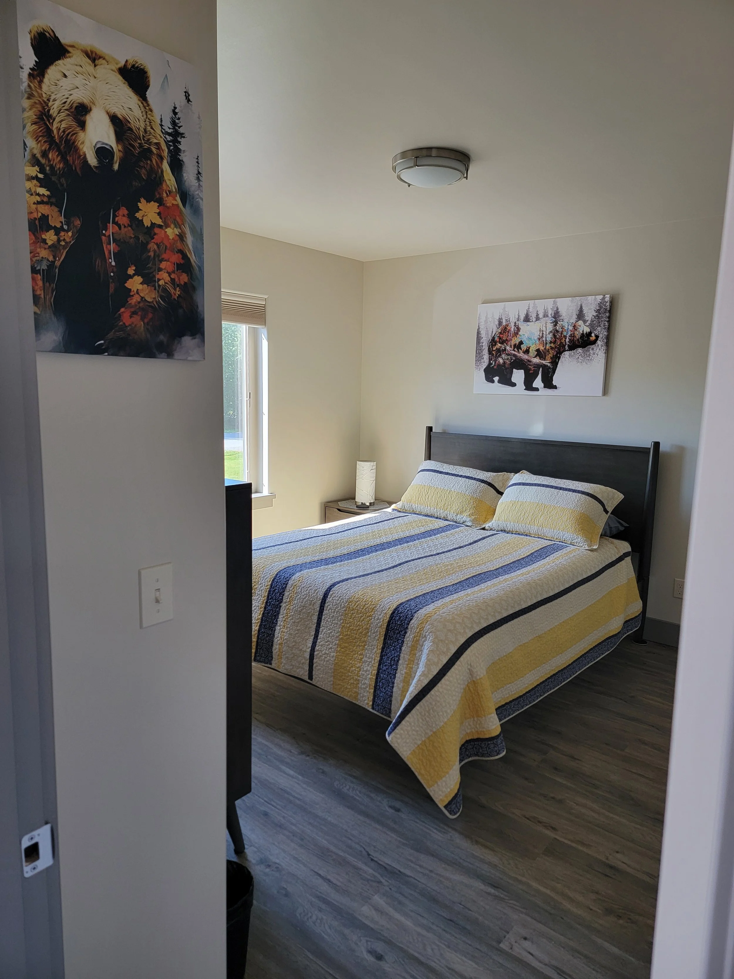 Cozy bedroom in a Lakeshore Lodging suite featuring a comfortable queen bed with a striped quilt, soft natural lighting, and bear-themed artwork for a charming Alaskan touch.