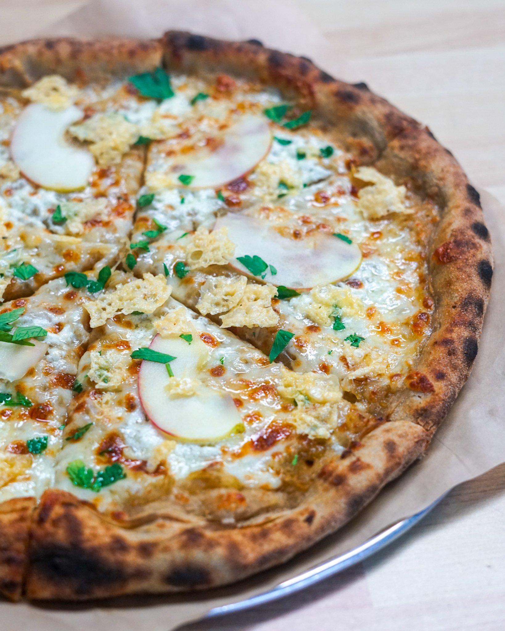Close-up of a pizza topped with apple slices, cheese, and crispy parmesan on a wooden table.