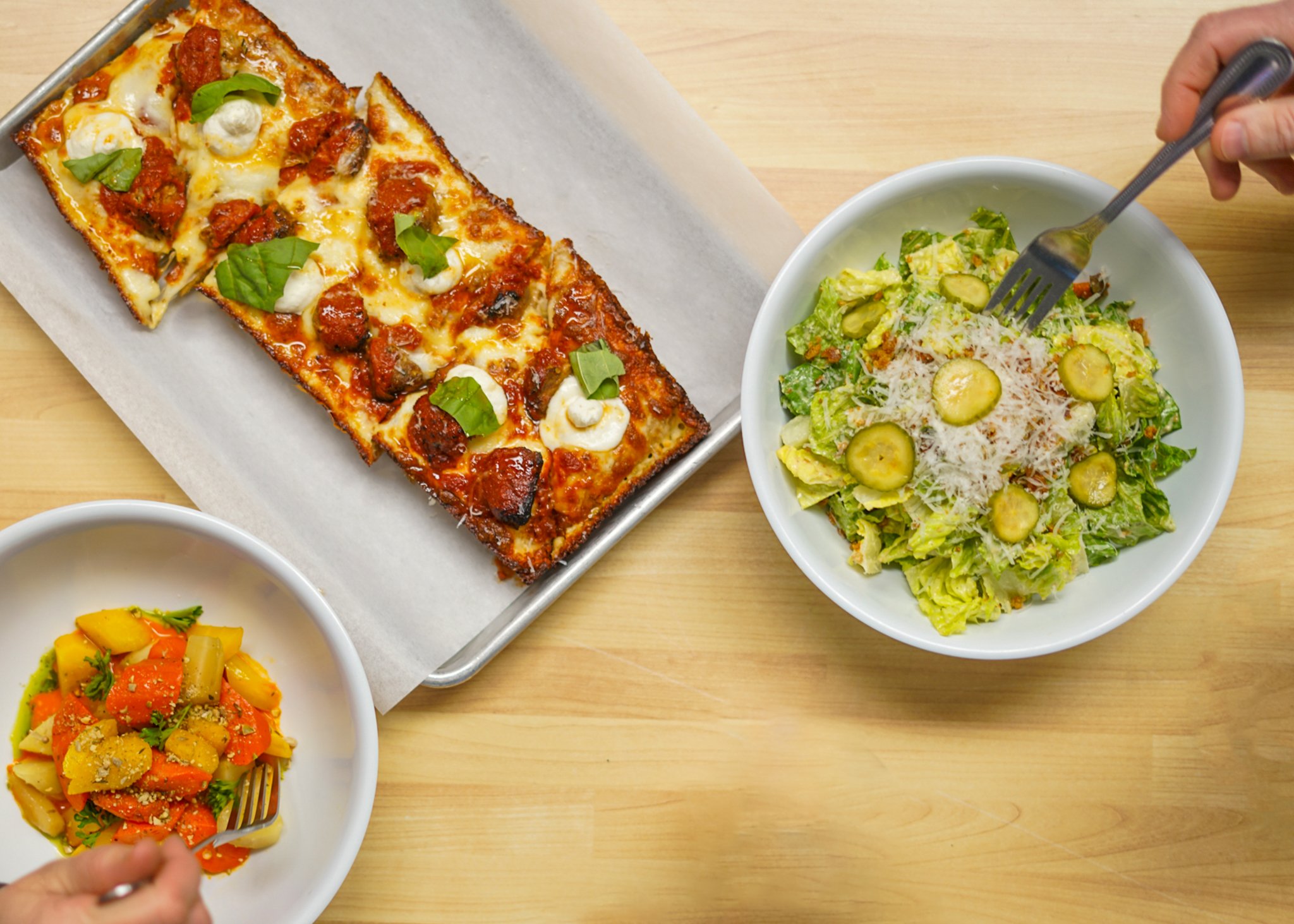 A rectangular pizza with cheese, meatballs, and basil on a tray, a bowl of Caesar salad with cucumbers, and a bowl of roasted carrots on a wooden table.