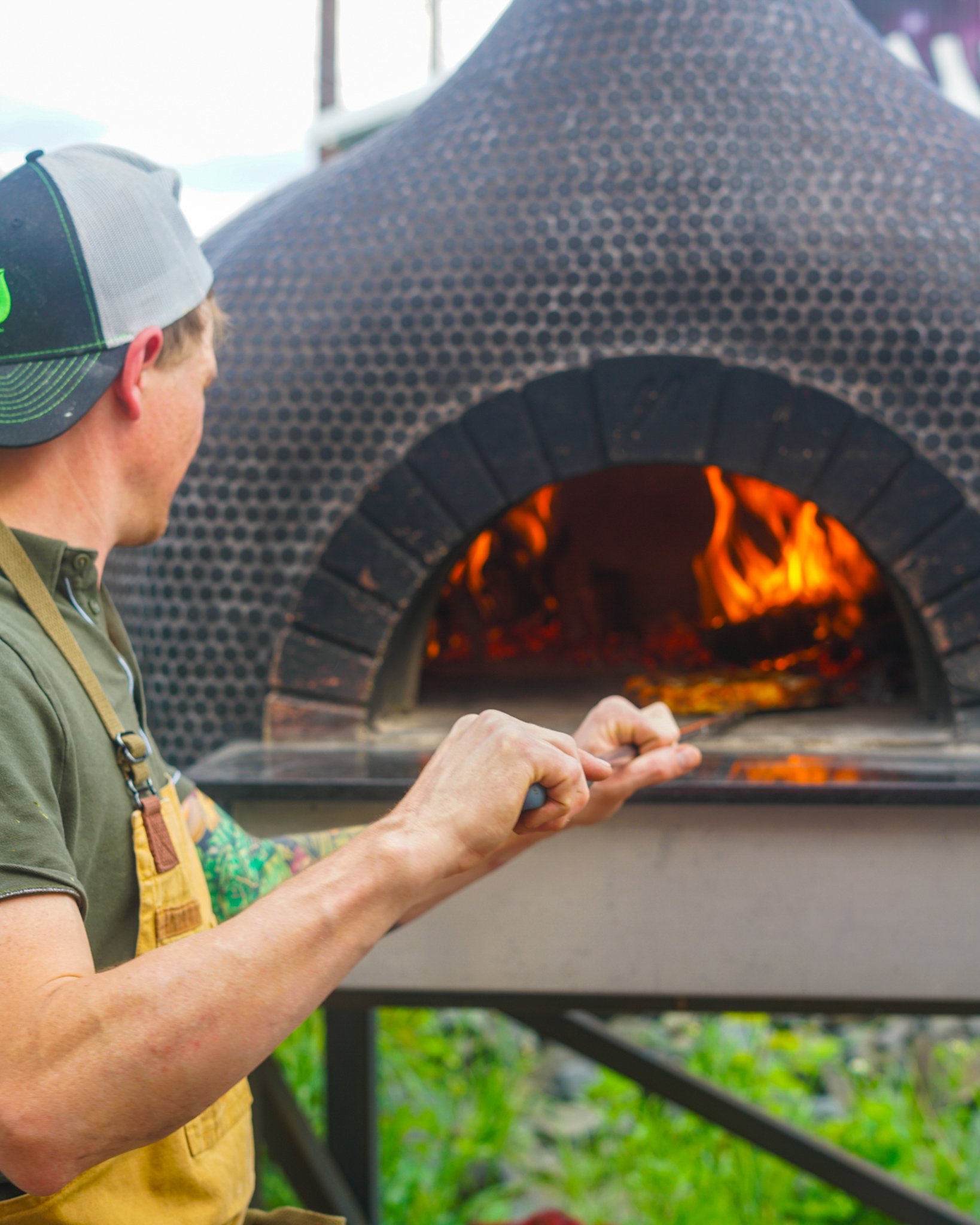 Person wearing a cap and apron baking pizza in a brick oven with flames inside.