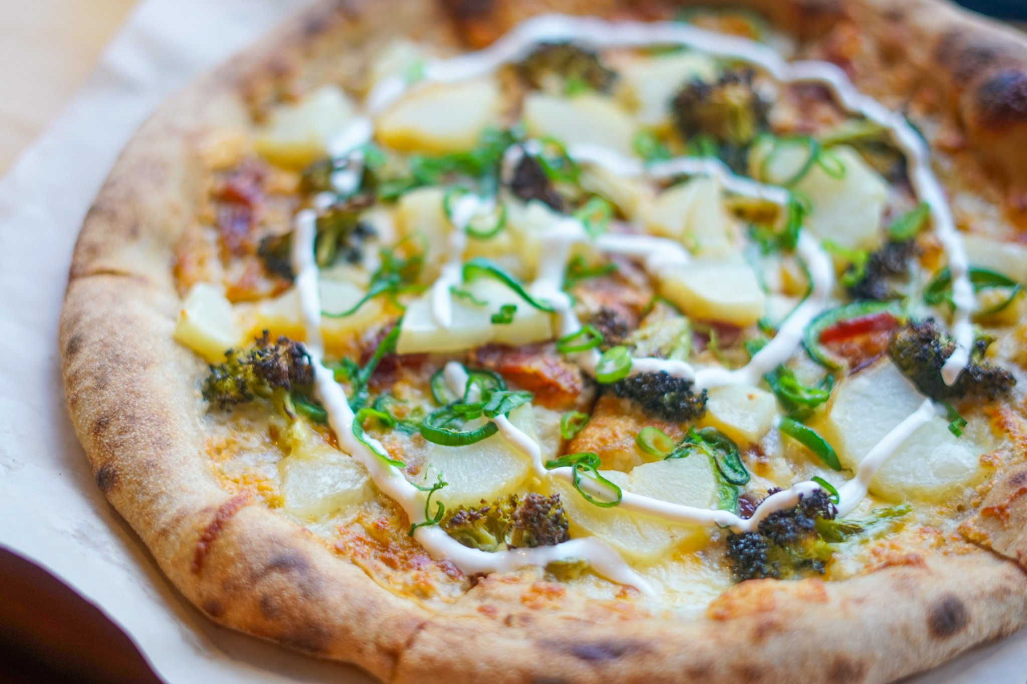 Close-up of a pizza with cheese, broccoli, potatoes, and white sauce drizzled on top.