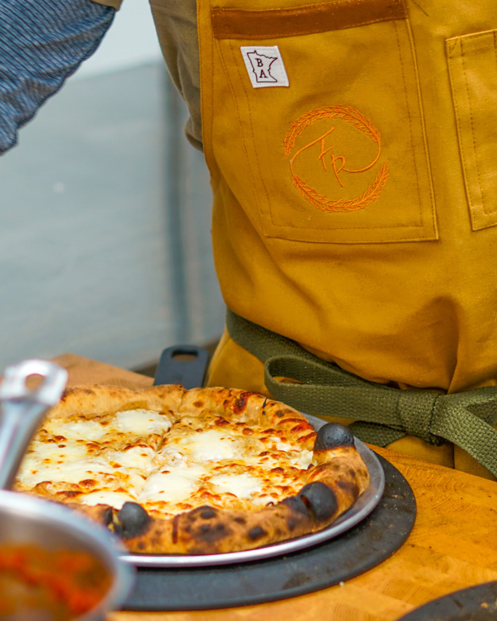 Close-up of a pizza with melted cheese on a tray, placed on a wooden table, with a person wearing an apron nearby.