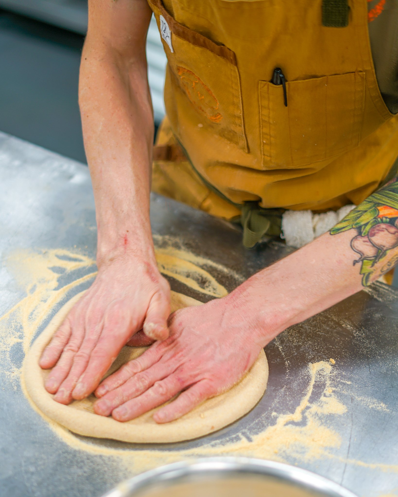 A person wearing an apron with tattoos on their arms is shaping pizza dough on a floured surface.