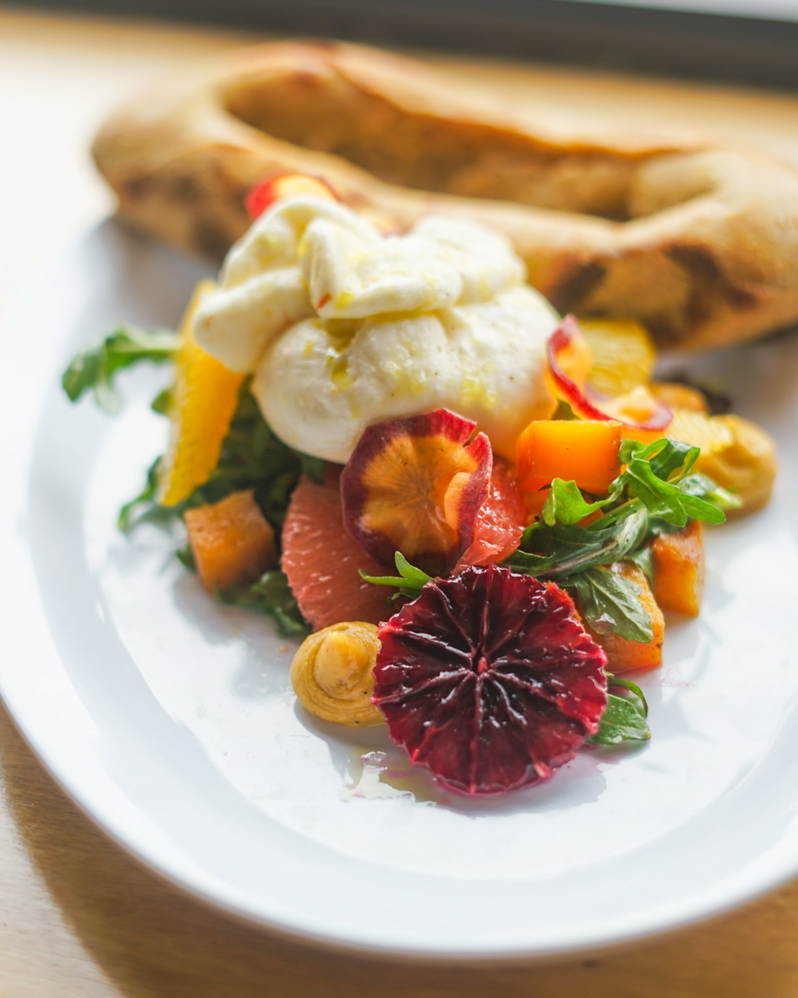 A plate of salad with mixed greens, sliced fruits, and burrata, accompanied by a bread roll in the background.