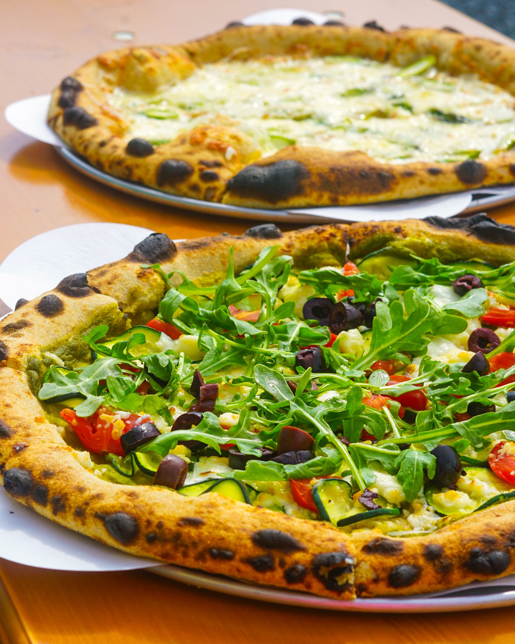Close-up of two pizzas on plates. The front pizza has arugula, cherry tomatoes, black olives, and melted cheese, while the back pizza is a cheese pizza with cheese and asparagus.