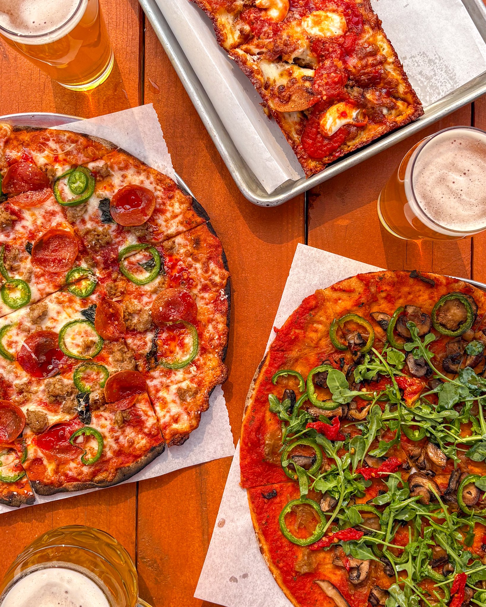 Three pizzas with toppings such as pepperoni, mushrooms, and arugula on a wooden table, with three glasses of beer.