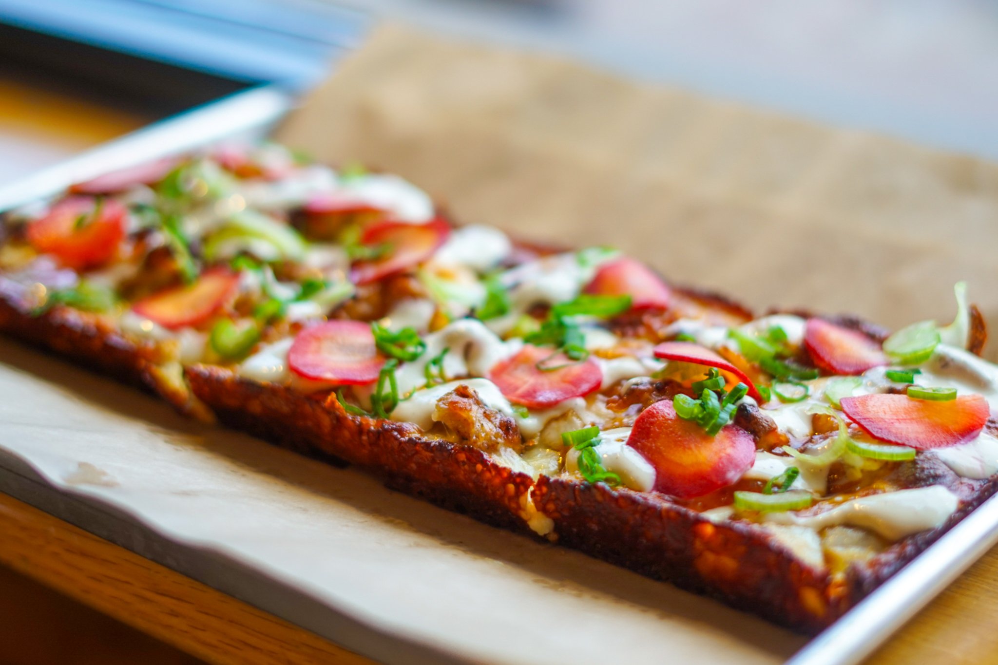 A rectangular pizza with a thick crust topped with cheese, sliced carrots, green onions, sitting on a tray with parchment paper.