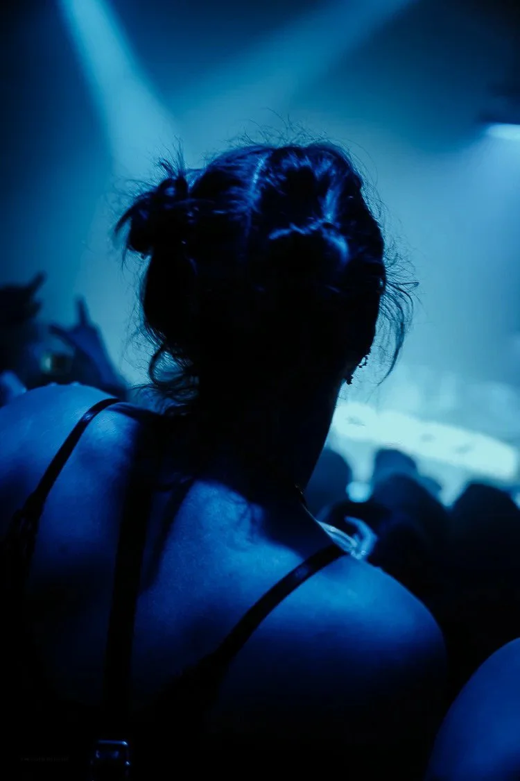 Back of a woman with dark hair in a messy bun, wearing a black strap top, in a dimly lit concert or club with blue lighting.