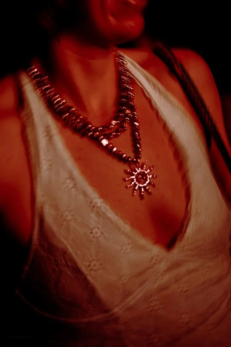 Close-up of a woman wearing layered silver necklaces, a white lace top, and a black strap, with dark background lighting.