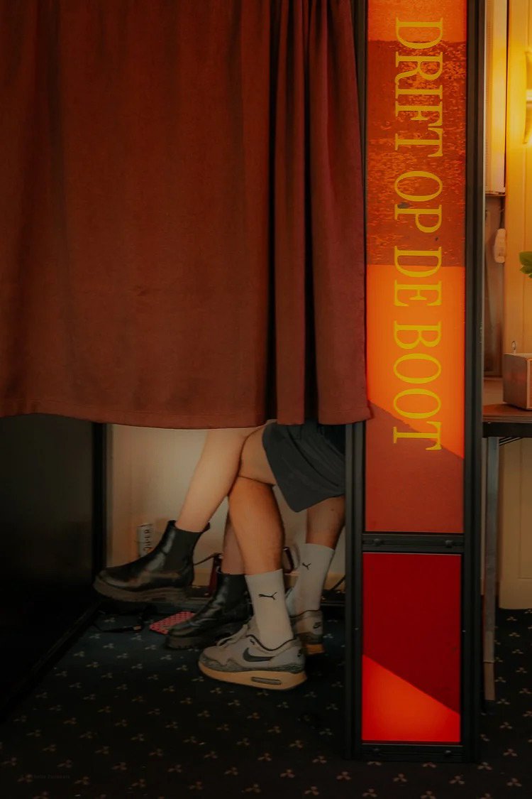 People sitting at a booth in a coffee shop, with their legs visible under a curtain, wearing sneakers and socks, near a sign that says "DREE OF BO".