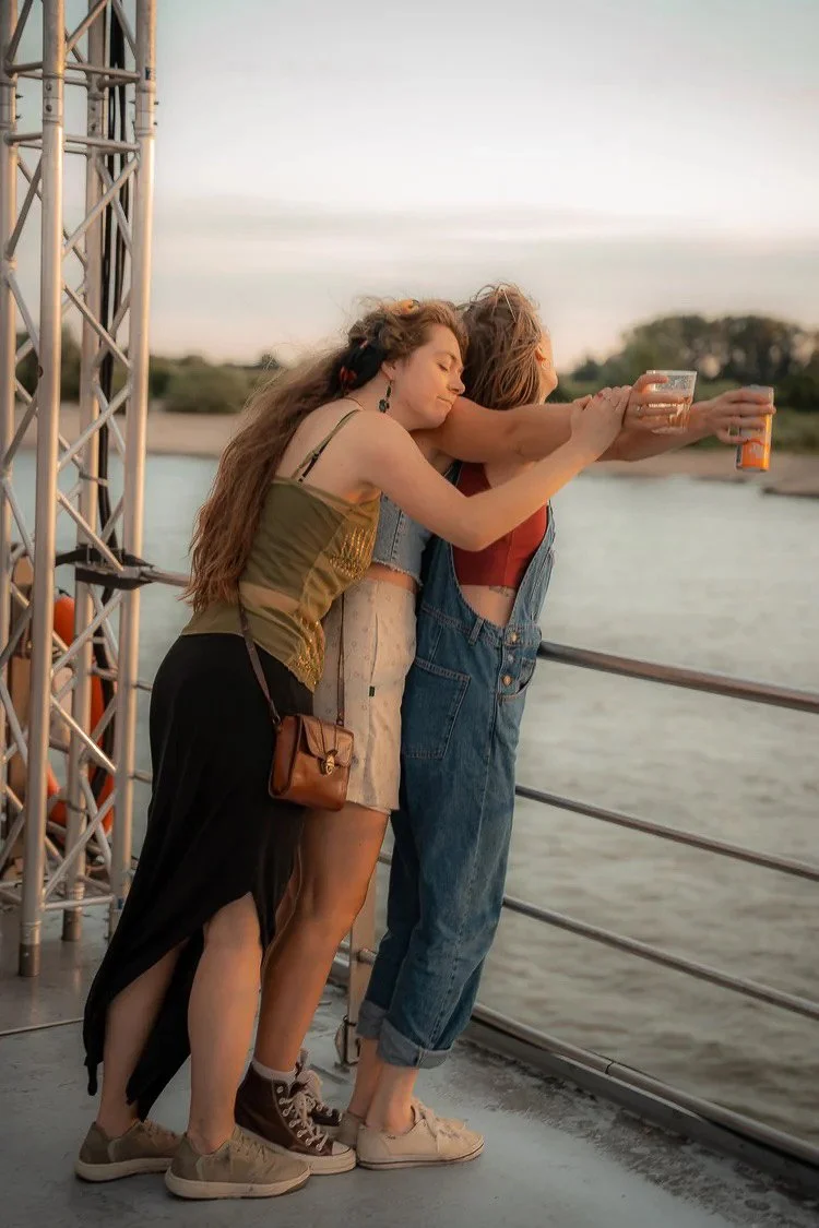 Three women standing closely together on a boat railing, taking a selfie with a water body and trees in the background during sunset.
