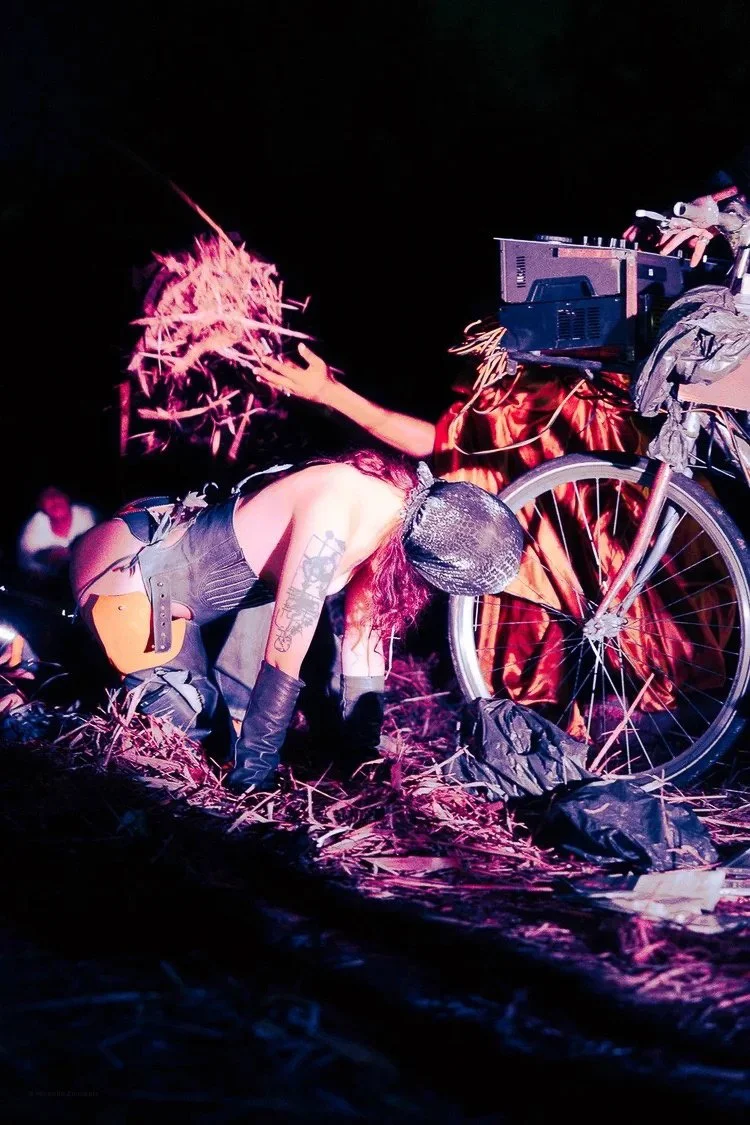 Person dressed in black with tattoos, wearing a helmet and gloves, kneeling on the ground near a bicycle, with a person standing behind, wearing a mask with a long nose, holding a lit cigarette; the scene appears theatrical or performance-based with 