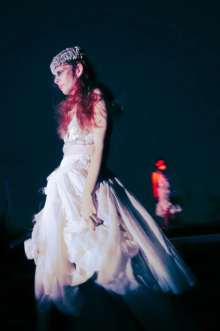 A woman in a white, flowing gown and a sparkling tiara walking on stage with a dark background, and a person in a hat seen in the background.
